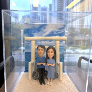 透明展示盒中的情侶人像公仔場景：米色砂地質感底座上，一座淺米色鳥居立於中央，後方為岩石與藍色海景、天空雲紋背板。左側角色穿藍色上衣與黑褲，右側角色穿藍灰色綁帶襯衫裙與白鞋，兩人面向前方、在鳥居前牽著彼此的手。展示盒置於木桌旁，窗外可見高樓與街景倒影。