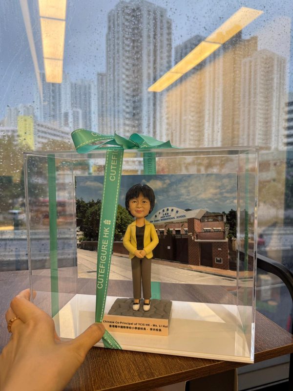 透明展示盒內的人像公仔站立於方形底座上：角色穿黃色西裝外套、黑色上衣與灰色長褲，雙手交疊於身前；身後放置學校外牆與校門的相片式背景。展示盒以綠色CUTEFIGURE HK緞帶綁結，前方名牌印有中英文職銜與名稱字樣；盒子置於木桌上，窗外雨滴與高樓景象映入玻璃。