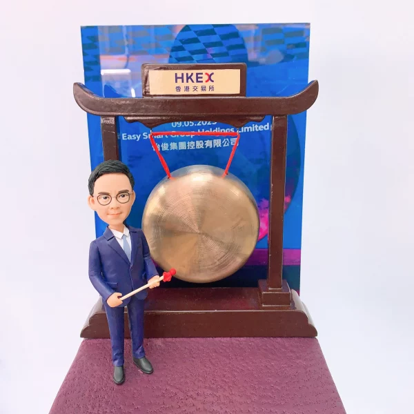 以香港交易所敲鑼儀式為主題的人像公仔：身穿深藍西裝與白襯衫的男性公仔，手持紅頭木槌站在銅鑼旁；銅鑼懸掛於深木色拱形架上，頂部銘牌印有「HKEX 香港交易所」。背板為藍色活動海報樣式，底座為暗紫色質感平台。整體呈現企業代表在港交所參與上市敲鑼的慶賀場景，作為金融領域的紀念人像公仔。