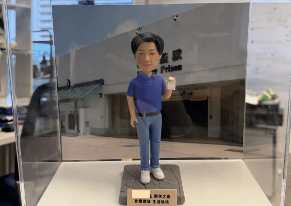 透明展示盒內的一體成形人像公仔：角色留短髮，穿藍色Polo衫、淡藍牛仔褲與白色運動鞋，黑色皮帶；右手抬起握著一張小卡或紙張，左手自然下垂。腳下是帶線條紋理的灰色方形底座。背板為某機構外牆與入口的照片式背景，可見英文單字 “Prison”。前方有淺金色小名牌，上面以黑色中文字寫著祝福語，包含「榮休之喜」「身體健康 生活愉快」等字樣。拍攝於白色檯面與室內環境。