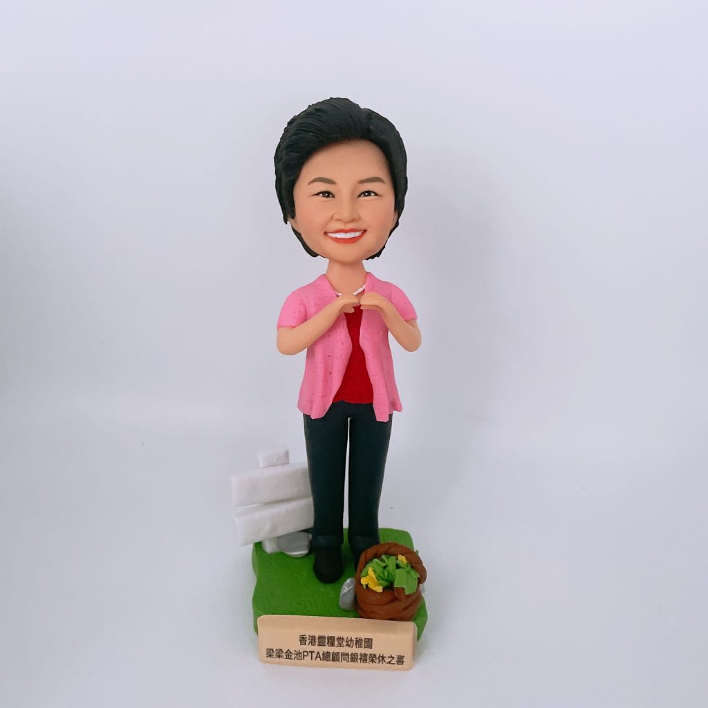 女士主題的人像公仔站在綠色草地質感的長方形底座上,短髮微笑,穿粉紅色開襟外套搭配紅色上衣、深色長褲與黑鞋;雙手於胸前比出愛心手勢。底座左後方有白色告示牌裝飾,右前方有裝著綠色蔬菜的小籃子;前置金色銘牌刻有「香港聖道堂幼稚園 路德金池PTA顧問榮休誌喜」等字樣。