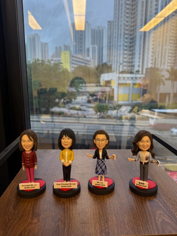 四款教師人像公仔排成一列放在木面桌上，背後是落地窗與高樓城市景。每個公仔立於黑紅圓形底座並配白色名牌：左起角色穿酒紅套裝；第二位穿黃色外套與深色褲；第三位戴眼鏡、深色外套搭配藍白格紋裙，手持小卡片；第四位長髮、穿灰色上衣配紅心別針並持卡片。名牌上以英中標示職銜與年份。整體呈現學校團隊展示氛圍。
