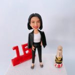 商務風格的人像公仔：一位黑色短髮女性面帶微笑，穿白色內搭、黑色修身長褲與黑色西裝外套（披肩式穿法），腳踩鑽飾開口高跟鞋，姿態自信、單手插袋。公仔站在白色紋理台座上；左側有大型紅色「15」立體數字與“Anniversary!”字樣，象徵十五週年里程碑；右側擺放香檳造型小物（奶油色瓶塞、黑黃環帶、底座上有紅色“J03”字樣）。背景為簡潔純白攝影棚。整體呈現企業高層／管理層慶祝周年的紀念人像公仔。