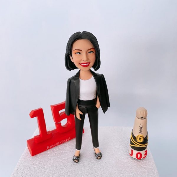 商務風格的人像公仔：一位黑色短髮女性面帶微笑，穿白色內搭、黑色修身長褲與黑色西裝外套（披肩式穿法），腳踩鑽飾開口高跟鞋，姿態自信、單手插袋。公仔站在白色紋理台座上；左側有大型紅色「15」立體數字與“Anniversary!”字樣，象徵十五週年里程碑；右側擺放香檳造型小物（奶油色瓶塞、黑黃環帶、底座上有紅色“J03”字樣）。背景為簡潔純白攝影棚。整體呈現企業高層／管理層慶祝周年的紀念人像公仔。