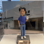 透明展示盒內的一體成形人像公仔：角色留短髮，穿藍色Polo衫、淡藍牛仔褲與白色運動鞋，黑色皮帶；右手抬起握著一張小卡或紙張，左手自然下垂。腳下是帶線條紋理的灰色方形底座。背板為某機構外牆與入口的照片式背景，可見英文單字 “Prison”。前方有淺金色小名牌，上面以黑色中文字寫著祝福語，包含「榮休之喜」「身體健康 生活愉快」等字樣。拍攝於白色檯面與室內環境。