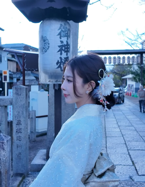 身穿淡色和服的女子側身站在石板路與神社風格街景前，髮髻上配有白色與粉色花飾與簪，背景見石燈籠與日文刻字。