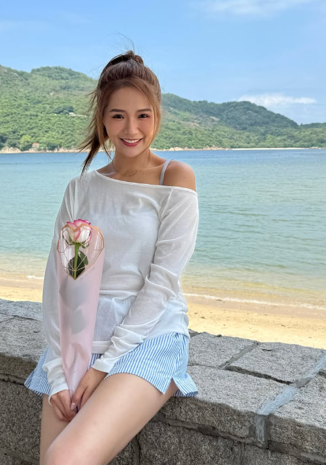 沙灘與海灣背景前，女子身穿白色上衣與藍白條紋短褲，手拿粉色包裝的玫瑰花。