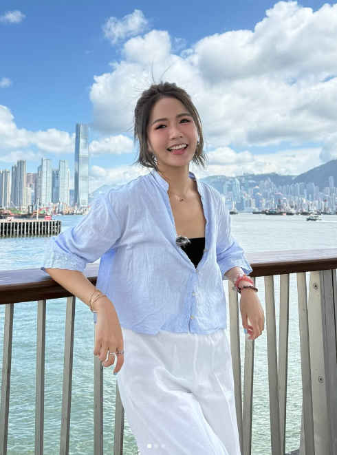 戶外海濱長廊，女子身穿淺藍襯衫與白色長褲，身後是藍天白雲與城市天際線。