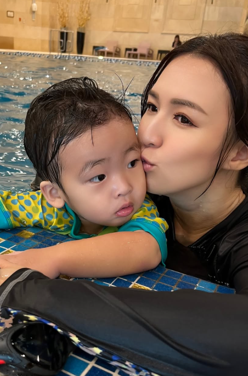 室內泳池邊，女子穿深色泳裝抱著幼童，臉貼向幼童臉頰。