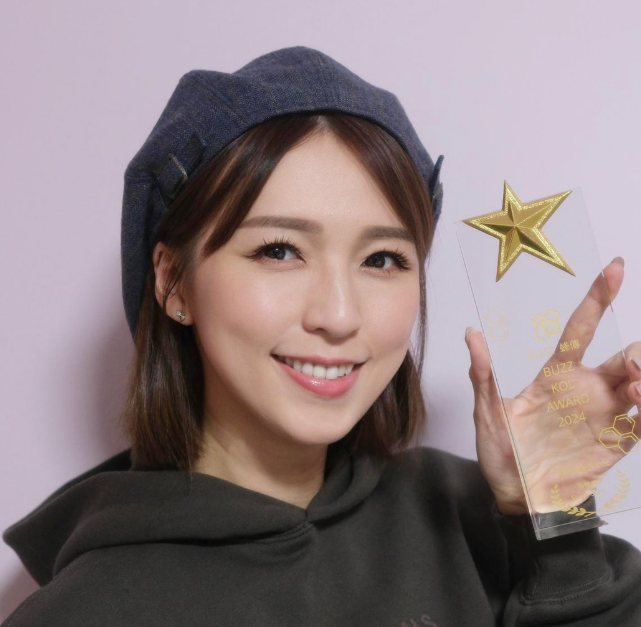 女子戴深色貝雷帽、穿深色連帽衛衣，手持透明獎座，獎座上有金色星形裝飾與英文字樣。