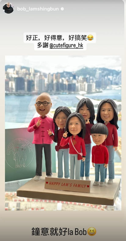 前景是一組家庭主題的人像公仔擺件：六位角色站在米色方形底座上，底座前有小牌寫「Happy Lam’s Family」。公仔皆穿紅色上衣與淺藍褲、白鞋，多人做出手指V手勢；其中左側一位戴黑框眼鏡、著粉紅外套。右前方擺放一張米白色祝賀卡，上書「Best Wishes」。背景為模糊的室內場景與坐著的成人身影。整體色調溫暖、桌面為木質。
