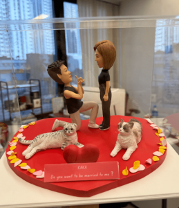 透明展示盒內的一組求婚情境人像公仔：紅色心形底座中央，一名短髮角色單膝下跪、手持戒指姿勢向對面短髮角色求婚，兩人皆穿黑色上衣與深色褲鞋。底座前方放置紅色立體愛心與粉色字牌，字牌上以白字寫著「KAKA Do you want to be married to me?」。左側有一隻灰白虎斑貓側躺，右側一隻白灰花貓坐姿，底座邊緣點綴粉、黃、白色花瓣與心形裝飾。背景是室內辦公環境與窗外城市高樓的景象。