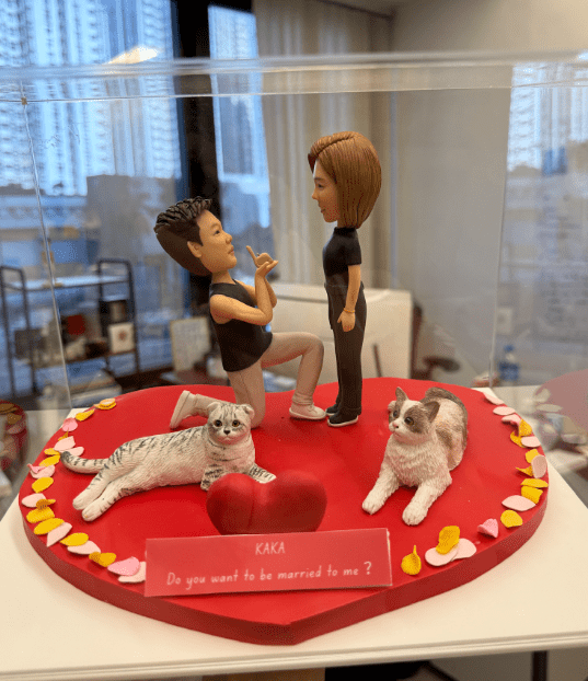 透明展示盒內的一組求婚情境人像公仔:紅色心形底座中央,一名短髮角色單膝下跪、手持戒指姿勢向對面短髮角色求婚,兩人皆穿黑色上衣與深色褲鞋。底座前方放置紅色立體愛心與粉色字牌,字牌上以白字寫著「KAKA Do you want to be married to me?」。左側有一隻灰白虎斑貓側躺,右側一隻白灰花貓坐姿,底座邊緣點綴粉、黃、白色花瓣與心形裝飾。背景是室內辦公環境與窗外城市高樓的景象。