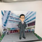 以校園／學術大樓為背景的展示作品：前景是一尊穿深灰色西裝、白襯衫與酒紅色領帶的人像公仔，佩戴黑框眼鏡、雙手插袋站立於灰色地磚地面。背景立牌印有一棟多層現代建築外觀，樓身窗框呈藍色，外牆可見「Academic Building」等英文字樣與校名標誌，入口處有拱形通道與紅色玻璃門。場景兩側以綠地與彩色小花點綴，呈迷你街景效果。