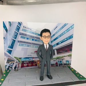 以校園／學術大樓為背景的展示作品：前景是一尊穿深灰色西裝、白襯衫與酒紅色領帶的人像公仔，佩戴黑框眼鏡、雙手插袋站立於灰色地磚地面。背景立牌印有一棟多層現代建築外觀，樓身窗框呈藍色，外牆可見「Academic Building」等英文字樣與校名標誌，入口處有拱形通道與紅色玻璃門。場景兩側以綠地與彩色小花點綴，呈迷你街景效果。
