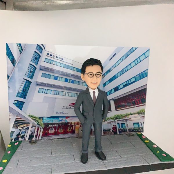 以校園／學術大樓為背景的展示作品：前景是一尊穿深灰色西裝、白襯衫與酒紅色領帶的人像公仔，佩戴黑框眼鏡、雙手插袋站立於灰色地磚地面。背景立牌印有一棟多層現代建築外觀，樓身窗框呈藍色，外牆可見「Academic Building」等英文字樣與校名標誌，入口處有拱形通道與紅色玻璃門。場景兩側以綠地與彩色小花點綴，呈迷你街景效果。