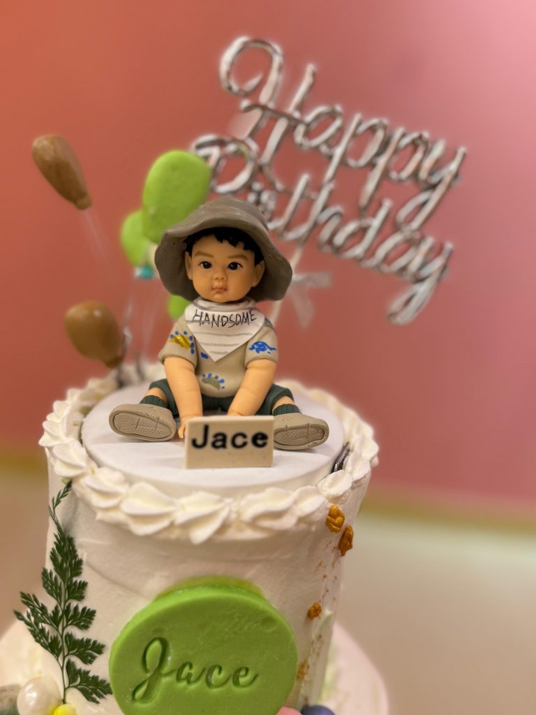 粉橘色背景下的奶油生日蛋糕特寫，蛋糕頂端坐著一個小朋友造型的人像公仔，戴寬邊帽、穿淺色上衣與短褲，圍兜寫有「HANDSOME」。公仔前方放著刻有「Jace」字樣的小姓名牌，後方有銀色「Happy Birthday」插牌與綠、棕色氣球裝飾；蛋糕側面貼有綠色圓形牌同樣壓印「Jace」，伴以葉片與彩色糖珠。