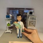 迷你場景中的醫生人像公仔站在灰色地台中央：穿淺藍手術服與白袍，雙手抱著裹綠毯的新生兒，右手持綠色復甦球。左後方是帶螢幕的生命監測儀與移動推車，右側為透明保溫箱模型；背景板呈現病房環境。前緣有淺膚色名牌，印有科室、姓名、職稱與年份字樣，呼應退休紀念主題。