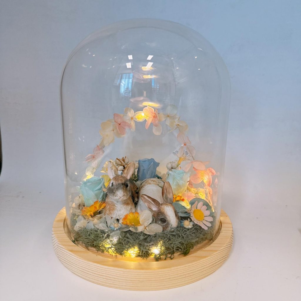 一座玻璃罩擺飾置於淺色木質圓形底座上，罩內鋪有苔蘚與碎石，中央有兩隻兔子動物公仔相依而坐；周圍點綴多朵永生花與小雛菊、藍色與粉色玫瑰，後方以粉橘色系花瓣組成半圓拱門。底座四周有暖黃小燈串發光，整體呈溫暖柔和的夜燈效果。