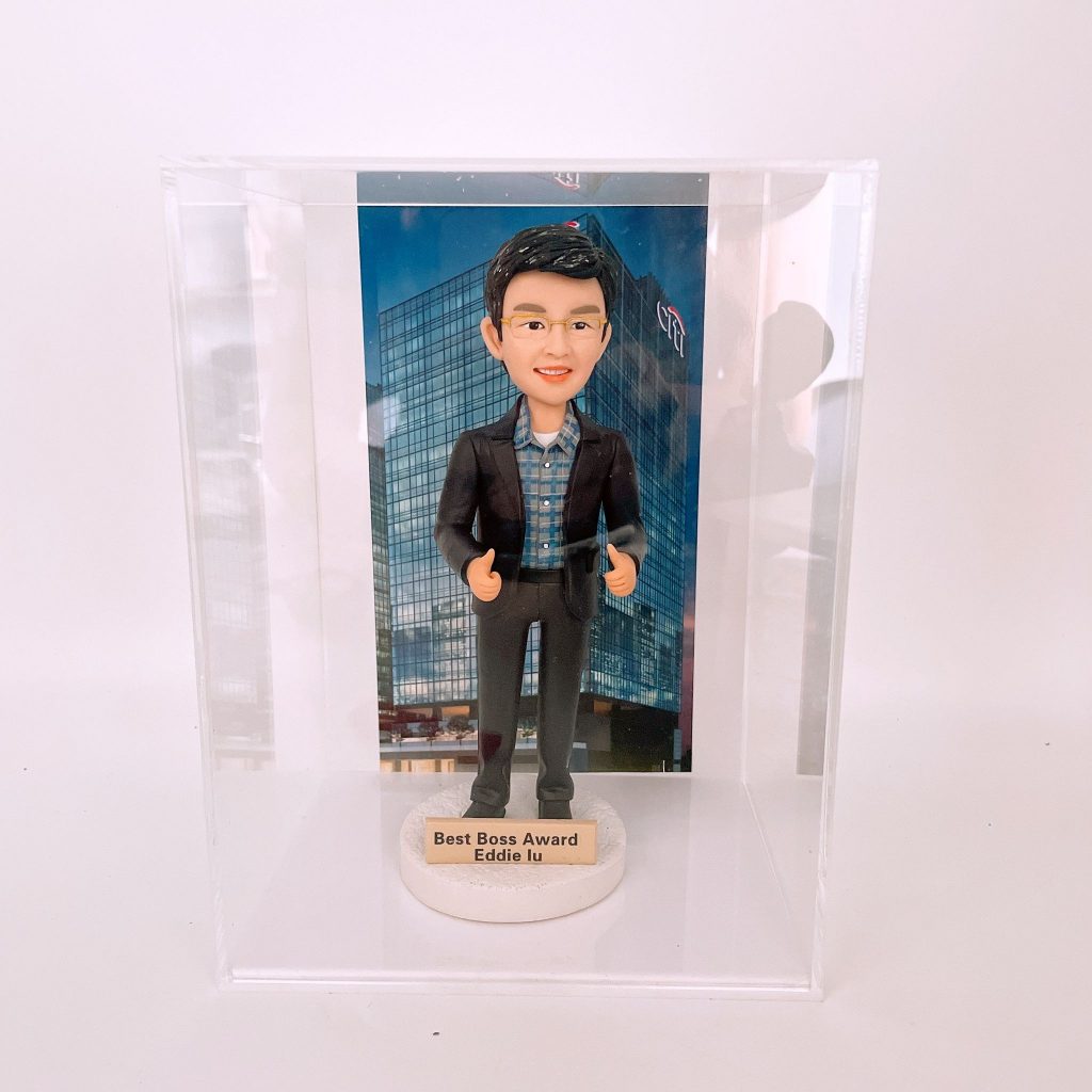 一個透明壓克力展示盒內的人像公仔：人物戴眼鏡、穿深色西裝與格紋襯衫，雙手比出大拇指站立於白色圓形台座上。台座前方貼有金色小銘牌，印有獎項與姓名字樣。公仔後方的背板為玻璃帷幕大樓的銀行總部外觀照片，整體擺放在白色背景前，呈現正式而典雅的禮品質感。
