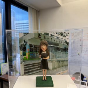 背後襯托著校舍照片,整體效果極具紀念意義。 公仔造型簡約典雅,身穿 黑色過膝連身裙,腳踏深色平底鞋,手中抱著書本,營造出 知性與專業 的氣質。細緻雕塑的臉龐帶著微笑,搭配標誌性的 及肩直髮與眼鏡造型,塑造出親切、可靠的形象。 底座以 綠色草地質感 呈現,呼應校園氛圍;透明保護盒讓公仔能被長久完整保存,防塵又具展示價值。這款設計特別適合作為 教師禮物、退休紀念、畢業送別或校園回憶收藏,帶有濃厚的紀念感與專屬心意。