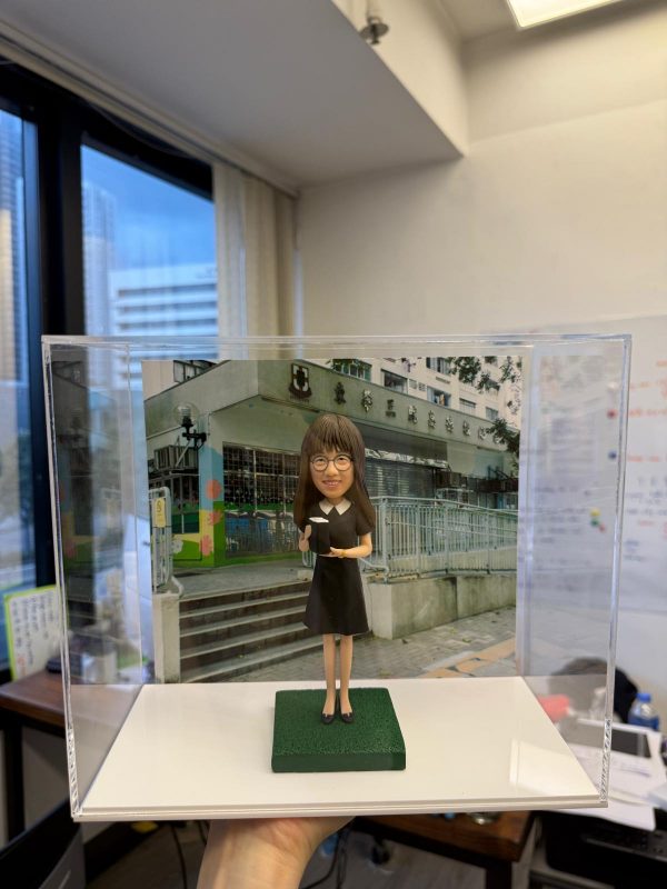 背後襯托著校舍照片，整體效果極具紀念意義。 公仔造型簡約典雅，身穿 黑色過膝連身裙，腳踏深色平底鞋，手中抱著書本，營造出 知性與專業 的氣質。細緻雕塑的臉龐帶著微笑，搭配標誌性的 及肩直髮與眼鏡造型，塑造出親切、可靠的形象。 底座以 綠色草地質感 呈現，呼應校園氛圍；透明保護盒讓公仔能被長久完整保存，防塵又具展示價值。這款設計特別適合作為 教師禮物、退休紀念、畢業送別或校園回憶收藏，帶有濃厚的紀念感與專屬心意。