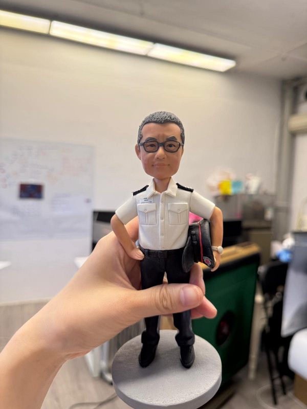 客製化個人職人公仔——制服警務人員/保安人員 手工人物公仔與細節 （圖片展示了一尊被手持著的客製化Q版軟陶公仔，主角是一位穿著制服的中年男性。公仔同樣採用頭大身小的Q版風格，面部特徵刻畫細膩，有著灰白的短髮和平頭造型，戴著黑框眼鏡，表情沈穩嚴肅，嘴角微微抿起。他身穿標準的白色短袖制服襯衫，肩上有肩章，胸前有口袋及名牌（依稀可見名字為「CHOW MING TAK」），下身穿著黑色長褲與黑皮鞋。他的姿勢是右手插腰，左手夾著一頂黑色大盤帽（帽沿有紅色線條裝飾），展現出執勤時的標準儀態。公仔固定在一個簡單的灰色圓形底座上。） 環境背景與光影 （這張照片是在室內拍攝的，背景是一間典型的辦公室或工作間。天花板上有明亮的長條形日光燈管，提供了均勻且充足的冷白光照明。背景處於失焦狀態（景深模糊），可以看到白板上貼有一些紙張或圖表，還有辦公桌、電腦螢幕以及雜物堆疊的角落，顯示出這是一個真實的工作環境。一隻手從左下方伸入鏡頭，穩穩地握住公仔的腿部及底座，將其展示在畫面中央。） 客製化公仔、Q版人像、制服公仔、警察/保安、退休禮物、榮休紀念、CHOW MING TAK、大盤帽、手工軟陶、專屬訂製 嚴肅專業、盡忠職守、正直穩重、職業榮譽、紀念價值、辦公室擺設、長官風範、制服細節
