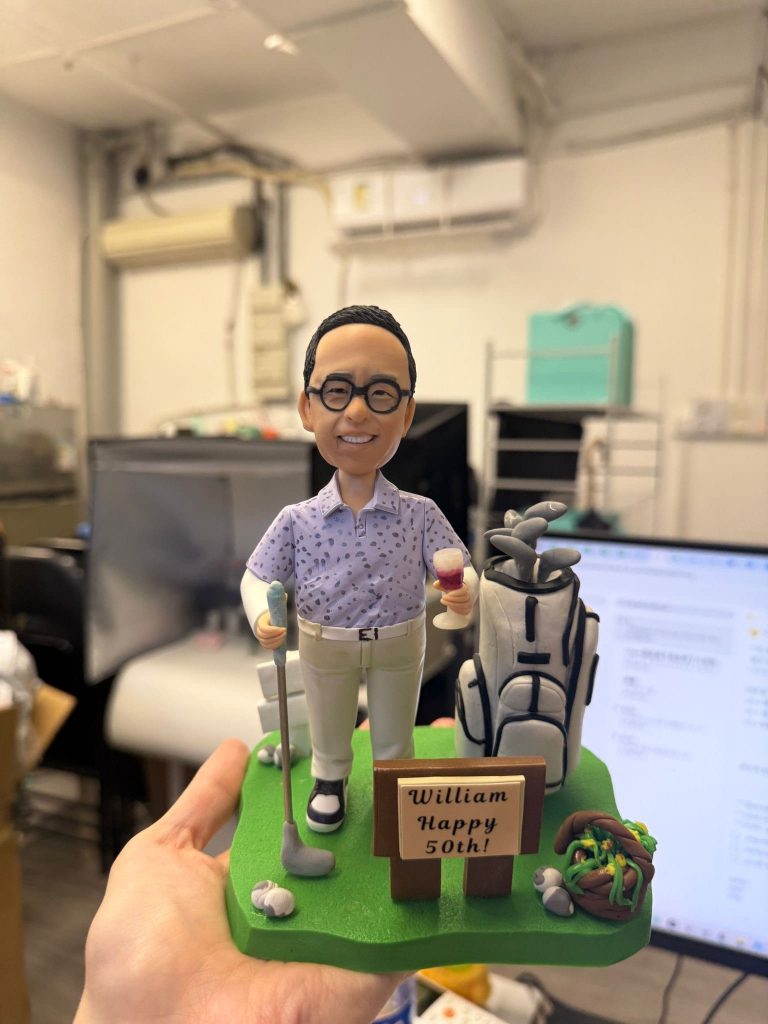 手掌托著一座高爾夫主題的人像公仔模型：人物短髮戴黑色圓框眼鏡，穿淺紫圖案襯衫與米色長褲、黑白球鞋，右手持酒杯、左手握高爾夫球桿，面帶微笑站在綠色草地台座上。人物旁有一個滿載球桿的高爾夫球包，地面散落幾顆高爾夫球與小石頭；前方立有木牌，上寫「William Happy 50th!」。台座一角有棕色藤圈與綠色裝飾。背景為室內工作環境，含電腦螢幕、空調與置物架等。