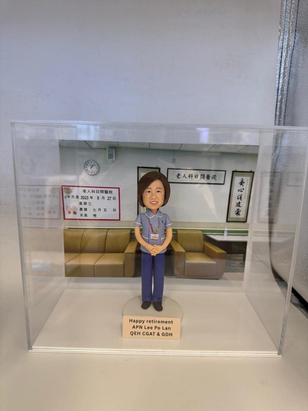 透明展示盒中的醫生人像公仔：角色穿藍灰色上衣與深藍長褲，掛識別證雙手於身前站立，置於淺色圓形底座。背板呈現門診等候區，有淺棕色沙發、牆上時鐘與中文標牌，左側公告板顯示日期與資訊。底座前方金色銘牌上寫有「Happy retirement APN Lee Po Lan QEH CGAT & GDH」。