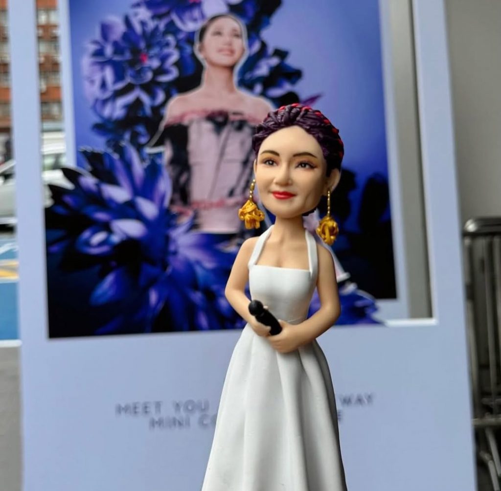 拼圖式畫面展示真人與對應的人像公仔造型對照：左側為一位穿白色長禮服、手持麥克風的舞台表演者，配戴誇張的金色耳飾與編髮造型；右側為同款元素的人像公仔，穿白色無袖長裙，手握黑色麥克風，戴金色耳飾與紫紅色編髮。其他照片中，公仔被置於海報前近拍，以及嵌入大束粉紫色系花束中央（玫瑰、馬蹄蓮、尤加利與葉材）作為花禮擺設，背景可見室外綠景。