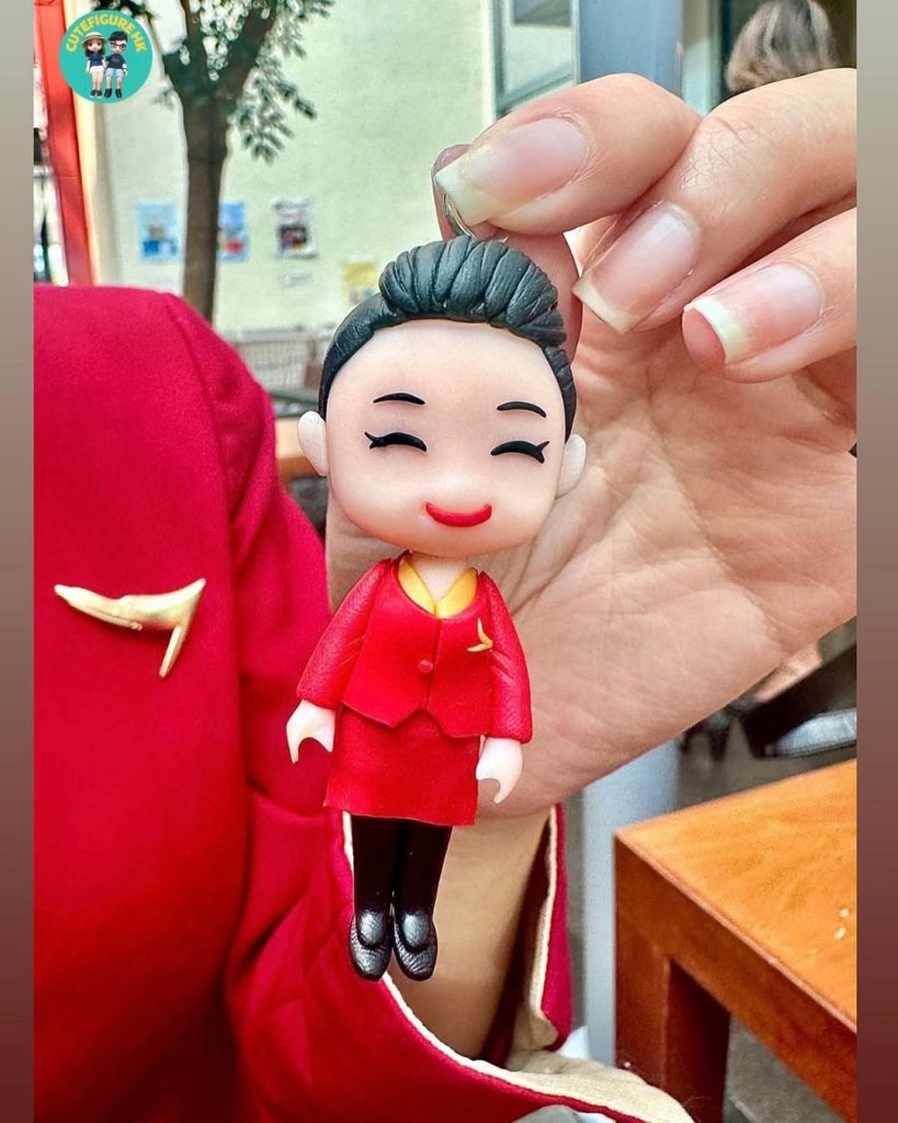 兩張照片組合：近拍中，一隻手以拇指與食指捏起一個Q版人像公仔吊飾。公仔為盤髮造型、閉眼微笑，穿紅色上衣與同色短裙套裝，內襯金色領口，胸前有金色徽飾，腳穿黑色鞋。背景可見穿著相同紅色制服的人士肩部與袖口。另一張為半身照，穿紅金配色制服的人士坐在桌邊，手持同款人像公仔欣賞，畫面角落附一個圓形小視窗再顯示公仔近景。