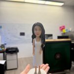 一款女性造型的人像公仔被手掌托起展示，站在淺灰圓形底座上；公仔留長黑色微捲髮，穿白色無袖中長洋裝與米色厚底涼鞋，五官精緻、姿態自然。背景為辦公室環境，遠處可見白板、文件、桌椅與大型綠色櫃體，景深虛化使公仔成為視覺焦點。
