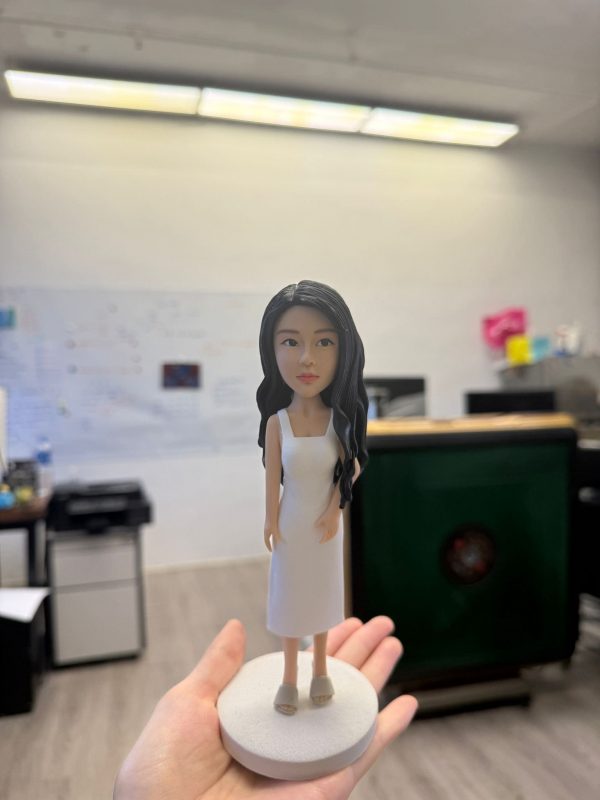 一款女性造型的人像公仔被手掌托起展示，站在淺灰圓形底座上；公仔留長黑色微捲髮，穿白色無袖中長洋裝與米色厚底涼鞋，五官精緻、姿態自然。背景為辦公室環境，遠處可見白板、文件、桌椅與大型綠色櫃體，景深虛化使公仔成為視覺焦點。