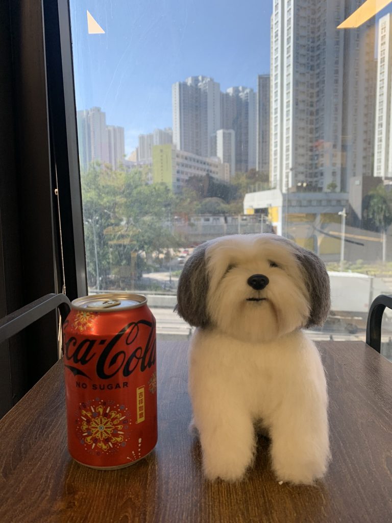 (客製化擬真羊毛氈西施犬公仔 手工寵物公仔細節（圖片右側展示了一隻極具真實感的羊毛氈狗狗公仔，外型特徵類似西施犬或拉薩犬；公仔全身覆蓋著蓬鬆柔軟的白色羊毛，雙耳則呈現灰黑色，毛流質感處理得非常細膩，還原了狗狗長毛的視覺效果；狗狗呈現乖巧的坐姿，眼神溫和，黑色的小鼻子與嘴巴線條栩栩如生；為了展示比例，公仔左側放置了一罐紅色的無糖可口可樂（Coca-Cola No Sugar），罐身印有「吉祥如意」與煙火圖案，公仔的高度略高於易開罐，顯示出其精巧的尺寸） 環境背景與光影（公仔與可樂罐置於一張深色木紋桌面上，背景是大片的落地窗景；窗外是晴朗的藍天與高聳密集的城市住宅大樓（類似香港的城市景觀），遠處還有綠樹與道路；明亮的自然光線透過窗戶照射進來，灑在羊毛氈公仔上，突顯了羊毛的柔和光澤與毛茸茸的立體質感，營造出悠閒明亮的午後氛圍） 羊毛氈、擬真寵物、西施犬、手工製作、客製化訂製、寵物紀念、可口可樂、比例尺、療癒系、毛小孩、仿真動物 寵物主人收藏、過世寵物紀念、擬真還原、生日禮物、居家擺飾、辦公桌小物、獨一無二的手作溫暖)