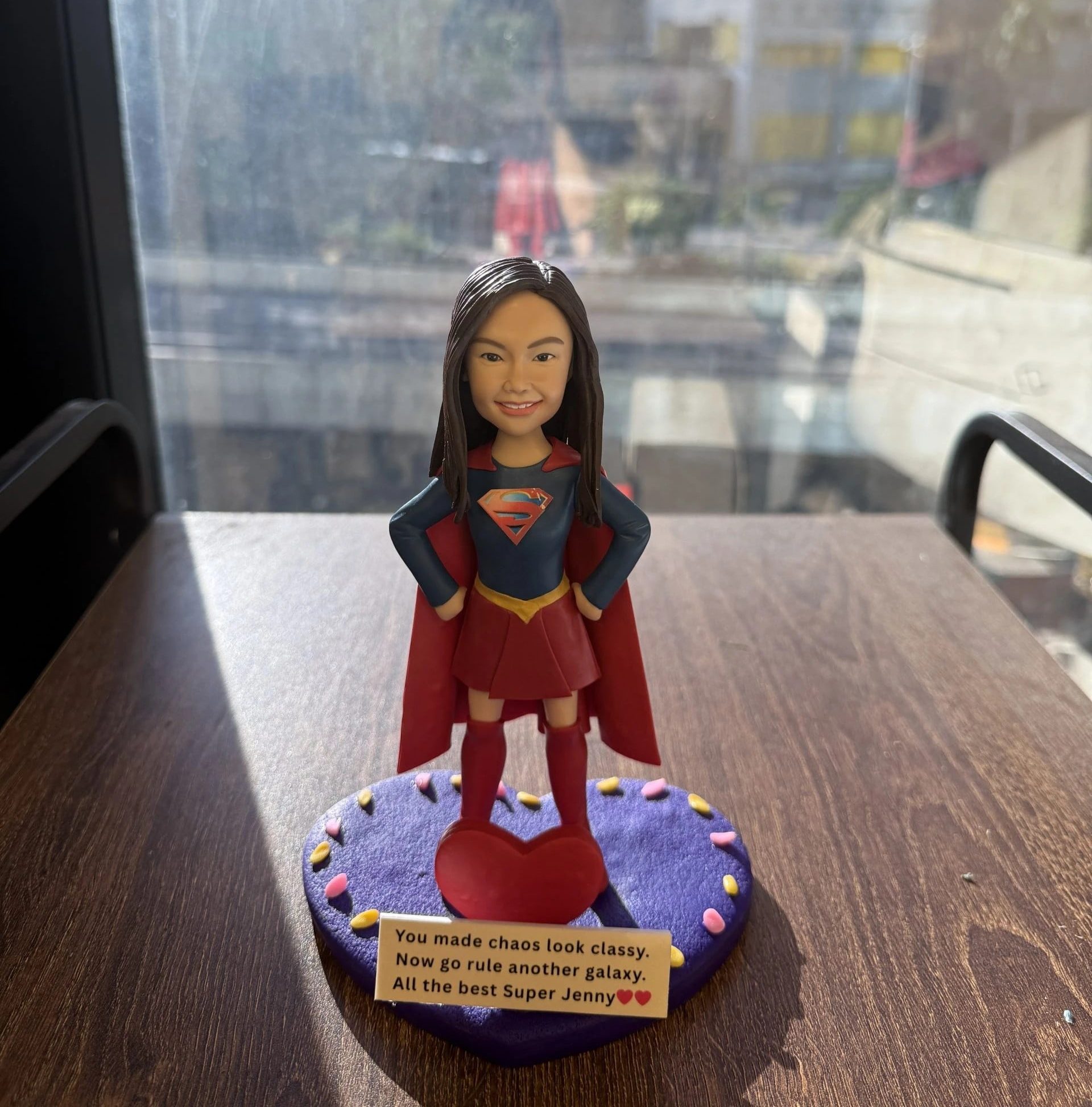 一尊以 女超人（Super Girl） 為主題的人像公仔作品。 中央公仔穿著紅色披風及深藍色戰衣，胸前綴有金紅色「S」標誌，擺出自信英勇姿勢。 面容細緻刻畫、笑容自然，五官比例與真人高度相似。 公仔立於一個紫色圓形底座上，邊緣裝飾以粉嫩心形點綴，前方放置紅色心形裝飾。鏡面背景映出窗外城市景觀，整體構圖充滿陽光光影與英氣氛圍。 作品象徵團隊中勇敢與靈感的精神，並作為送別同事的 Farewell紀念公仔。