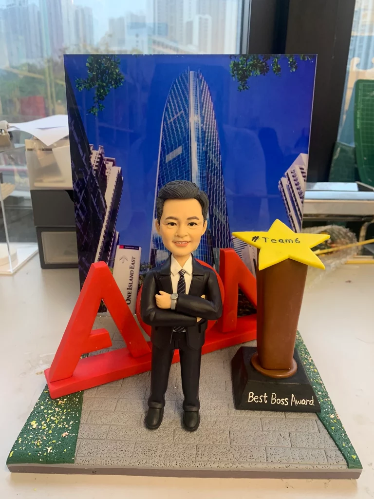一組桌面擺設：前景是一位穿深色西裝、打條紋領帶的人像公仔，雙臂交叉站在磚紋灰色台面上；左後方有紅色立體「AON」字母結構，右側是一座棕色獎座上方的黃色星星，上寫「#Team6」，黑色底座以白字標註「Best Boss Award」。背景板為藍天下的玻璃幕牆摩天大樓與街景，底座兩側撒有金色碎片點綴。