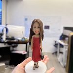 一隻精緻的人像公仔站在淺灰圓形底座上，被手掌托起展示；公仔為長直髮女性造型，穿紅色細肩帶連身裙與黑色厚底高跟鞋，右手拎著黑色小包。背景是開放式辦公室，可見桌面、螢幕、白板與文件，景深虛化讓主體更突出。
