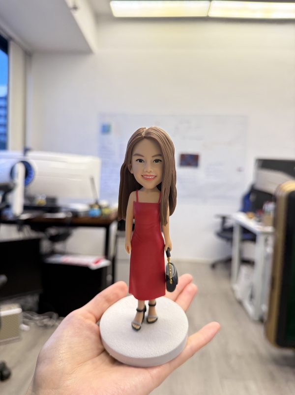 一隻精緻的人像公仔站在淺灰圓形底座上，被手掌托起展示；公仔為長直髮女性造型，穿紅色細肩帶連身裙與黑色厚底高跟鞋，右手拎著黑色小包。背景是開放式辦公室，可見桌面、螢幕、白板與文件，景深虛化讓主體更突出。