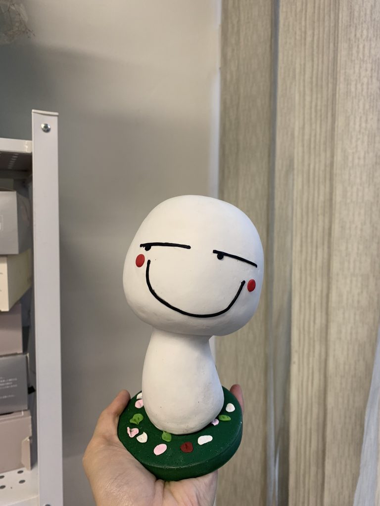 白色人形公仔，擁有卡通化的大頭和瞇眼笑容，臉頰兩邊有紅色腮紅，站在綠色圓形底座上，是訂造卡通人物公仔的趣味作品。