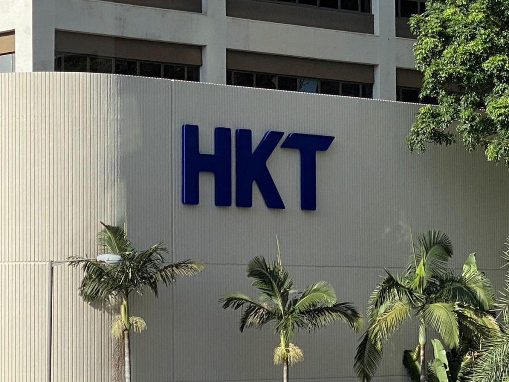 一棟淺色建築物外牆上掛著藍色「HKT」企業標誌，前方有幾棵棕櫚樹，象徵著企業形象與榮譽的企業紀念品。