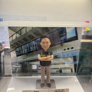 一款人像公仔置於透明展示盒內，背景為機場航班資訊顯示板與候機大廳結構。公仔穿深色背心式制服與卡其色長褲，胸前有徽章與「CUSTOMS」字樣臂章，雙臂交叉站立於灰色底座。展示盒前方放有名牌與獎牌式小牌座，顯示職稱與表揚訊息，整體呈現祝賀升遷的紀念氛圍。