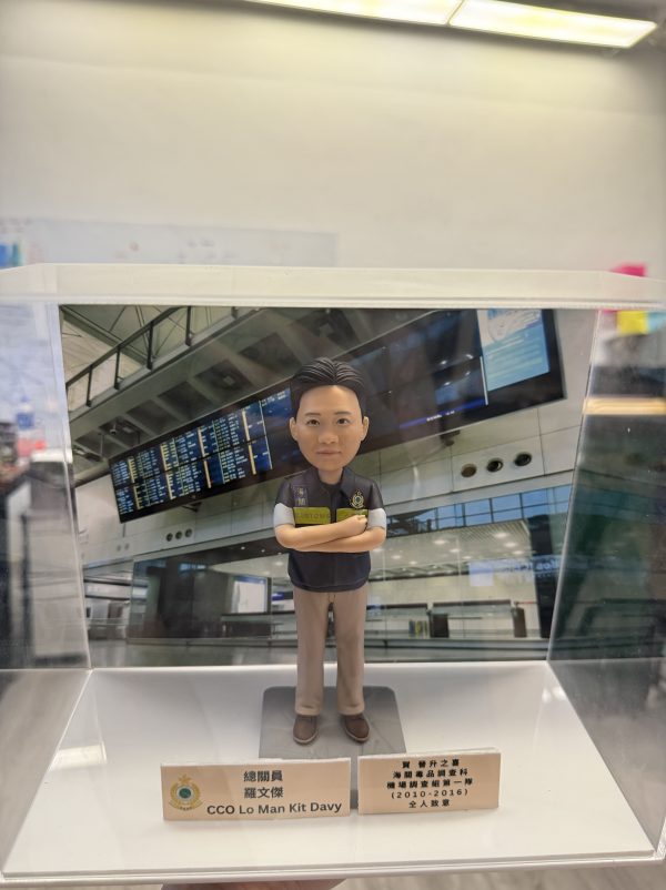 一款人像公仔置於透明展示盒內，背景為機場航班資訊顯示板與候機大廳結構。公仔穿深色背心式制服與卡其色長褲，胸前有徽章與「CUSTOMS」字樣臂章，雙臂交叉站立於灰色底座。展示盒前方放有名牌與獎牌式小牌座，顯示職稱與表揚訊息，整體呈現祝賀升遷的紀念氛圍。