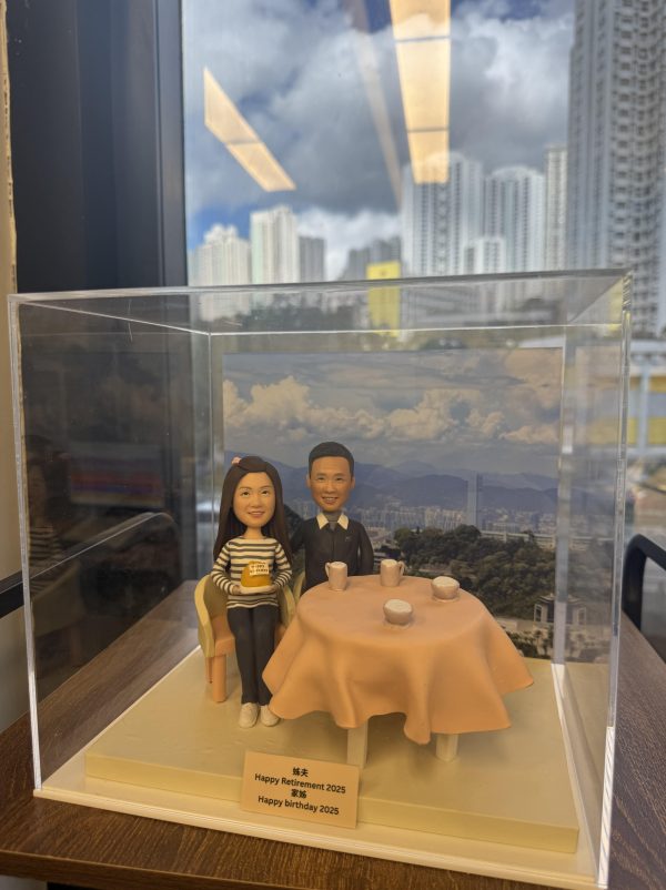 透明壓克力展示盒中的雙人退休人像公仔：左側角色穿條紋上衣、黑褲與白鞋，雙手捧杯入席；右側角色著深色西裝，並肩坐在圓桌旁。桌面覆粉橘色桌布，擺放三只茶杯與一只小盅，呈現酒樓用茶氣氛。前方小銘牌寫有「Happy Retirement 2025」與「Happy birthday 2025」。背板為城市高樓與遠山景致，整體置於木質檯面。