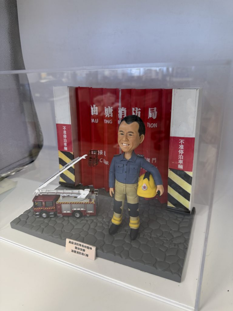 透明展示盒內的人像公仔場景：一位消防主題公仔站在灰色六角石紋台座上，身穿深藍上衣與卡其色防護褲、黑靴與護膝，雙腿綁有黃色反光帶；左臂抱著黃色消防頭盔。背景是紅色消防局摺疊車庫門與黑黃斜紋防撞柱，門上可見白色「消防局／FIRE STATION」字樣。公仔右前方擺放一輛紅色雲梯消防車模型，台座前角有淺色小銘牌，上印黑字祝賀訊息。整體置於透明壓克力盒中。