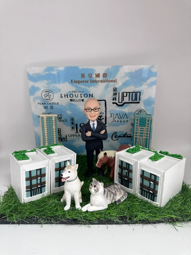 立體場景擺設：草地底座上擺放三棟白色獨立洋房模型，屋頂與草皮綠化細節可見；中央站著一位穿深藍西裝、雙手交叉的人像公仔，身後有迷你小馬模型；前景有一隻白色狗與灰白虎斑貓並列而坐。背板印有「Emperor International 英皇國際」及多個樓盤名稱與簡化建築圖示。