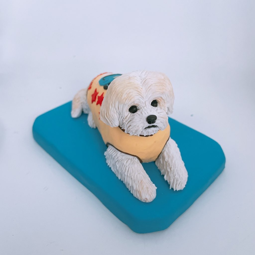 (客製化黏土手作馬爾濟斯公仔 手工寵物公仔細節（圖片展示了一隻精緻的手工黏土狗狗公仔，外型特徵為馬爾濟斯或比熊犬；公仔呈現趴臥姿勢，全身毛髮以細膩的刻痕技法表現出蓬鬆的層次感，毛色主要為白色，耳朵部分帶有淡淡的奶油色漸層；狗狗身穿一件黃色的寵物衣，背部裝飾有紅色星星圖案與藍色細節；五官刻畫生動，黑亮圓潤的眼睛與小鼻子展現出無辜可愛的神情；公仔被固定在一個圓角的長方形藍色底座上，整體造型小巧精緻） 環境背景與光影（公仔置於純白色的背景中進行拍攝，畫面乾淨簡約，沒有多餘的雜物干擾視覺焦點；光線均勻柔和，類似攝影棚的柔光箱效果，消除了強烈的陰影，清楚呈現出黏土材質的紋理細節與色彩飽和度，凸顯出手作藝品的質感） 輕黏土、軟陶、馬爾濟斯、比熊犬、手工模型、客製化訂製、寵物公仔、Q版造型、立體擺飾、手作禮物、仿真動物 寵物紀念品、車內擺飾、辦公桌療癒小物、送給狗主人的禮物、獨一無二的手捏溫度、收藏品)