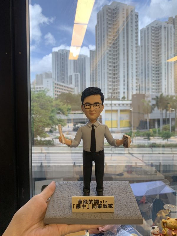(客製化Q版人物公仔——專業男教師手持文具紀念擺飾 手工人物公仔細節（圖片展示了一尊客製化的Q版男性人物黏土公仔，主角戴著黑框眼鏡，身穿淺藍色長袖襯衫並打著深色領帶，下身搭配黑色西裝褲與皮鞋，展現專業俐落的形象。公仔雙手張開，右手持有一把長尺，左手拿著一台計算機，暗示其可能為數學或理科教師。公仔站立於一個仿水泥質感的灰色方形底座上，底座前方貼有一塊金色名牌，黑字寫著「萬能的譚Sir 『羅中』同事致敬」，表達了同事間的敬意與讚賞） 環境背景與光影（公仔被一隻手托舉在空中，背景是一扇大落地窗。透過窗戶可以看到戶外清晰的城市景觀，包括高聳的白色住宅大樓、藍天白雲以及下方的街道與綠樹。室內天花板的長條形燈光倒映在玻璃窗上。自然光線從窗外透入，使公仔的輪廓清晰，面部表情溫和自然，背景的城市景觀為公仔提供了現代感的襯托） 軟陶公仔、真人公仔訂製、Q版人像、數學老師禮物、理科老師、教師節禮物、手工捏塑、客製化名牌、職業公仔、手持文具 同事送禮、榮休紀念、感謝師恩、辦公室擺設、獨家訂製紀念品、校園紀念、活動贈禮)