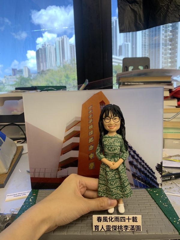(客製化Q版女教師公仔——校園背景榮休紀念擺飾 手工人物公仔細節（圖片展示了一尊客製化的Q版女性人物黏土公仔，主角留著黑色長捲髮並有瀏海，戴著黑框眼鏡，面帶親切微笑。她身穿一件綠色底白色花紋的長袖連身裙，腳踩白色休閒鞋，雙手優雅地交疊在身前。公仔最顯著的特點是其背景板，是一張「樂善堂梁植偉紀念中學」橙色校舍大樓的實景照片，還原了校園場景。公仔立於仿灰色磚石紋路的底座上，前方名牌寫著「春風化雨四十載 育人靈傑桃李滿園」，點明了這是一份紀念執教四十週年的榮休或長期服務禮物） 環境背景與光影（公仔被放置在一個看似工作檯的環境中，由一隻手扶持展示。背景稍顯雜亂，可見堆疊的書籍、文件及製作工具，暗示這是在製作完成後的檢視環節。後方是大片落地窗，窗外是藍天白雲與高樓大廈的城市景觀，明亮的自然光線照射進來，使公仔的色彩鮮豔，綠色裙裝與橙色校舍背景形成鮮明對比） 軟陶公仔、真人公仔訂製、Q版人像、女教師禮物、校長禮物、榮休紀念、校園場景還原、綠色碎花裙、樂善堂梁植偉紀念中學、長期服務獎 感謝師恩、四十週年紀念、教育工作者、獨家訂製、辦公室擺設、移居紀念、桃李滿門、手工藝品)