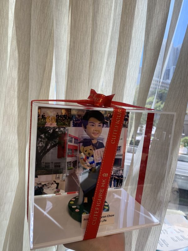 一座透明禮盒包裝的桌面公仔擺設，外繫紅色緞帶與金色字樣。盒內中央是一位短髮微笑的人像公仔，穿深色禮袍式披肩與長裙，雙手捧著泰迪熊與一張迷你證書/卡片；腳邊是綠色圓形草地底座，散落白粉色小花。背景板拼貼多張校園相關照片，包含紅白相間外牆的校舍、校園樹蔭、辦公室場景與團體合照。底部白色標牌印有英文名稱與“Principal”字樣。整體放置在手掌上，背後是室內米色窗簾與窗外城市景致。