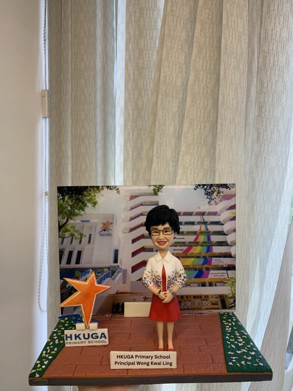 手托一座小型桌面場景模型：中央是一位短髮、戴眼鏡的人像公仔，穿白色外套搭配彩色花卉印花袖口與紅色連身裙，雙手於腹前交握站立。背景立牌印有一所學校建築照片，立面帶有紅色水平線與彩虹色斜紋裝飾，四周樹葉遮蔭。左前方立著一顆橙色星形標誌與「HKUGA PRIMARY SCHOOL」字樣的小牌。底座為紅棕色磚紋地面，兩側綠色帶狀區域點綴白色小花；前方金屬銘牌寫著「HKUGA Primary School Principal Wong Kwai Ling」。